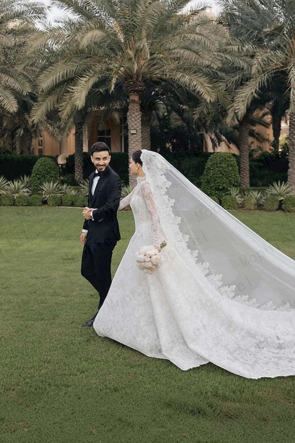 TheBlissWedding white lace long sleeve A-line wedding dress with a sweep train worn by a bride walking with a groom.
