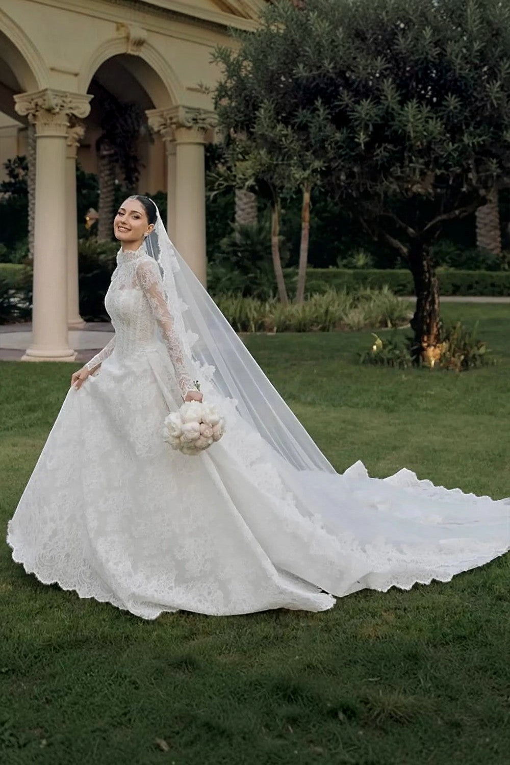 TheBlissWedding white lace A-line wedding dress with long sleeves, high neck, and a dramatic sweep train in a garden.