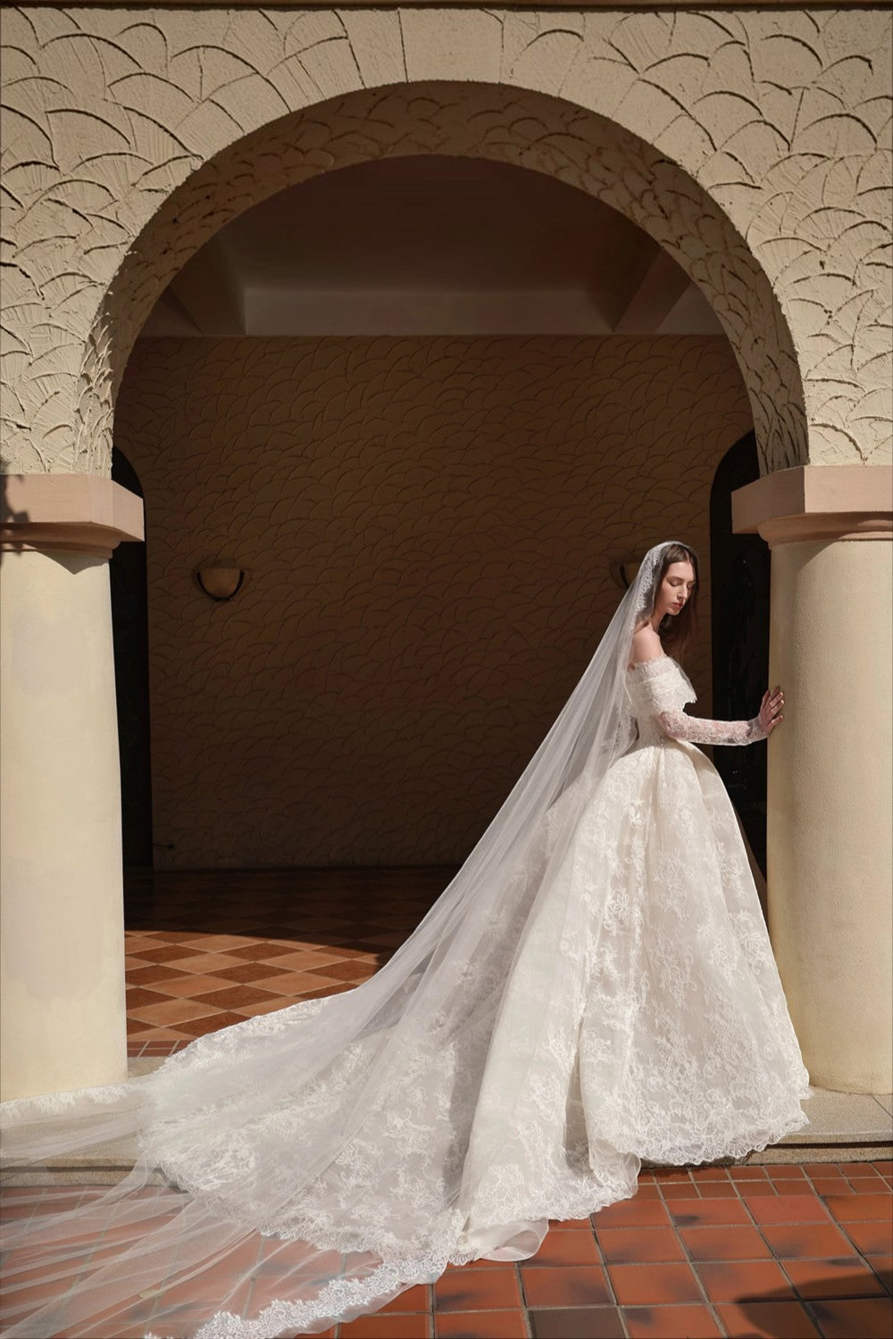 TheBlissWedding white off-shoulder A-line wedding dress with long lace sleeves and a long veil in a stone archway