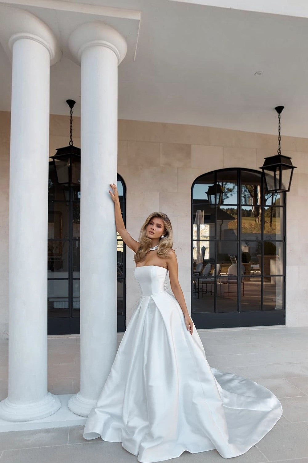 TheBlissWedding strapless white satin A-line wedding dress with a sweep train, worn by a model against white pillars.