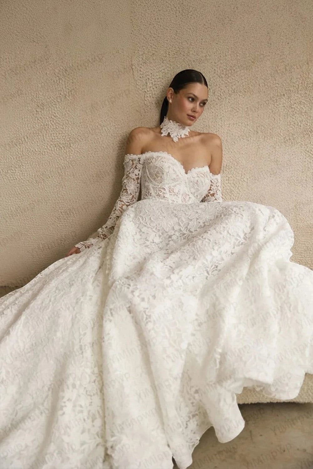 TheBlissWedding white lace off shoulder wedding dress with long sleeves and a sweetheart neckline on a seated model.